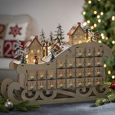 a wooden calendar with houses and christmas trees in front of a lit up christmas tree