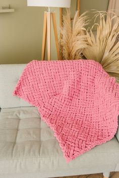 a pink knitted blanket sitting on top of a couch next to a vase with dry grass