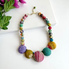 a necklace made out of different colored stones and beads on a white surface with flowers in the background