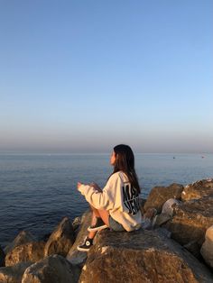 a woman sitting on top of a rock next to the ocean looking out at the water