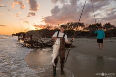 a man standing on the beach holding a fish and fishing rod with another person in the background