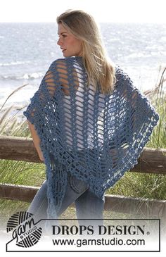 a woman wearing a blue crocheted shawl standing next to a fence with the ocean in the background