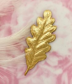 a gold leaf brooch sitting on top of a pink and white background with feathers