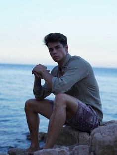 a man sitting on top of a rock next to the ocean with his feet crossed