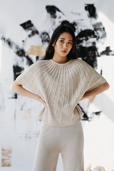 a woman standing in front of a white wall wearing a sweater and wide legged pants