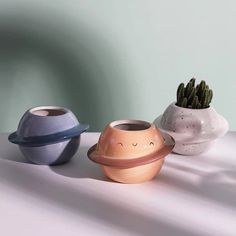 three ceramic pots with plants in them on a white tablecloth, one is shaped like saturn and the other has a face drawn on it