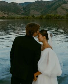 a bride and groom standing in front of a lake with mountains in the background on their wedding day