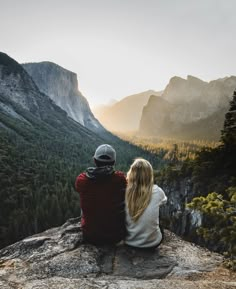 two people sitting on top of a cliff looking at the mountains and trees in the distance