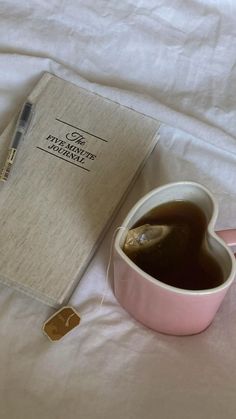 a cup of coffee next to a book and pen on a white bed with sheets