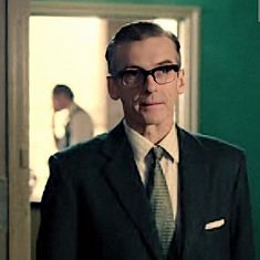 a man wearing glasses and a suit in front of a green wall with an open door