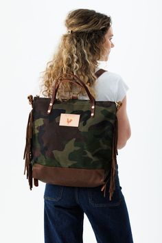 Woman wearing the camouflage bag with the meanwhile logo and brown fringe on her back with backpack straps. Waxed Canvas Backpack With Waxed Finish, Waxed Canvas Satchel Backpack With Waxed Finish, Brown Waxed Canvas Standard Backpack, Waxed Canvas Backpack With Leather Backing, Brown Waxed Canvas Leather Backpack With Adjustable Strap, Brown Leather Backpack With Adjustable Strap And Waxed Canvas, Brown Waxed Canvas Backpack, Chic Diaper Bag, Chicken Logo