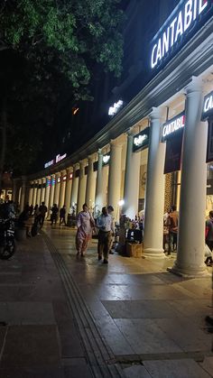 people are walking around in front of a store at night with lights on and columns lit up