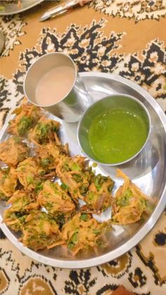 a silver plate topped with food next to two bowls filled with green liquid and sauce