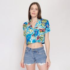 Vintage 80s cropped button up blouse in blue with a Hawaiian-themed print of palm trees, volcanos, and flowers. Measurements and Condition:    Fits like: Labeled women's medium Fabric: Rayon - labeled machine wash cold Brand: Andrew Sport, made in USA Condition: Excellent, appears to have been hemmed up by a previous owner.   Length: 15.5" Chest: 39" Waist: 38" Shoulders (seam to seam): 16" Sleeve: 6.5" Shown on a 5'9" model with measurements of 33"-24"-37", usually wears size XS to small. See our FAQ for more info on sizing and condition ratings. Retro Short Sleeve Blouse For Beach, Retro Short Sleeve Blouse For Vacation, Summer Hawaiian Blouse With Tropical Print, Hawaiian Style Summer Blouse With Tropical Print, Summer Hawaiian Tropical Print Blouse, Hawaiian Style Summer Tropical Print Blouse, Blue Short Sleeve Hawaiian Shirt For Vacation, Blue Fitted Hawaiian Shirt For Beach, Fitted Blue Hawaiian Shirt For Summer