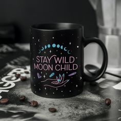 a black coffee mug with the words stay wild moon child on it next to some coffee beans