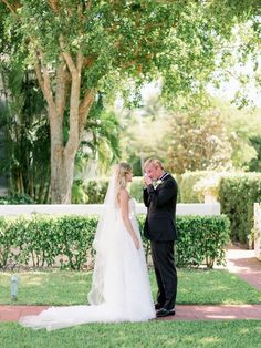 a bride and groom standing in front of a tree on their wedding day, looking at each other