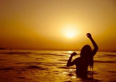 a woman standing in the ocean at sunset raising her arms up to the sun above her head