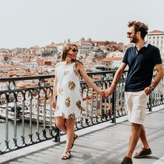 a man and woman holding hands while walking on a bridge over water with buildings in the background