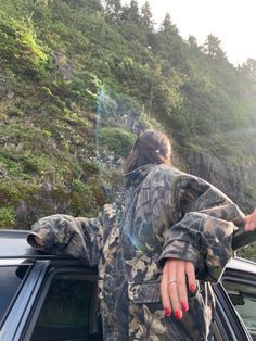 a woman standing next to a car in front of a mountain with trees on it