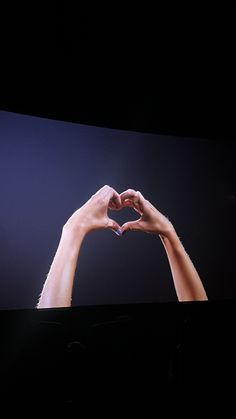 two hands making a heart shape with their fingers on a large screen in the dark