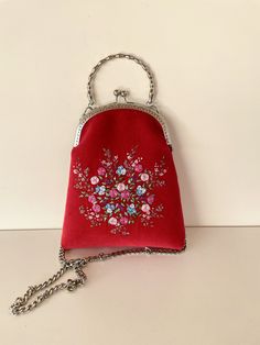 a red purse with floral embroidered on the front and chain at the bottom, sitting on a white surface