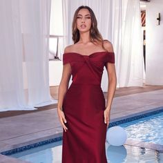 a woman standing in front of a swimming pool wearing a red dress with off the shoulder sleeves