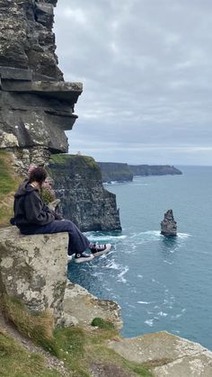 a man sitting on top of a cliff next to the ocean with cliffs in the background