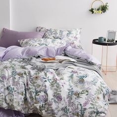 a bed with purple sheets and pillows in a white room next to a side table