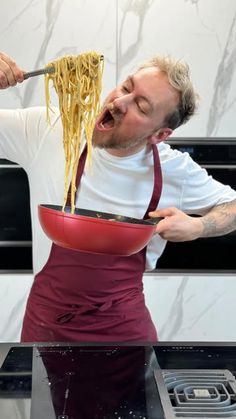 a man in an apron is eating spaghetti from a red bowl on top of a stove