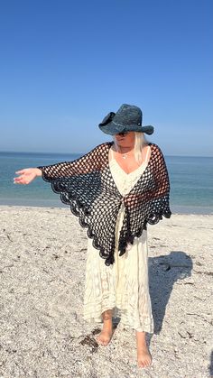 a woman standing on the beach wearing a hat and shawl over her shoulders, with her arms outstretched