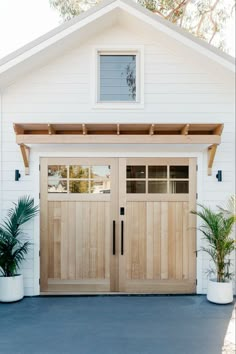 a white house with two wooden garage doors and plants in pots on the front porch