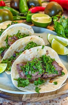 three steak tacos on a plate with limes and cilantro