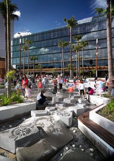 many people are walking around in front of a large glass building with palm trees on both sides