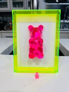 a pink teddy bear in a green frame on a counter top with other items behind it