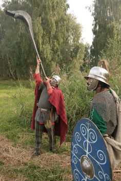 two men dressed in medieval costumes and helmets, one holding a spear while the other holds a shield