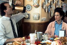 two people sitting at a table with food and drinks in front of the man drinking from a coffee mug