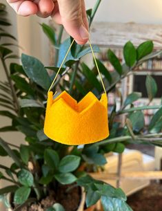 a hand is holding a yellow felt crown ornament in front of a potted plant