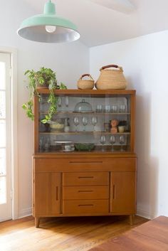 a wooden china cabinet with glass doors and plants on top, in a white walled room