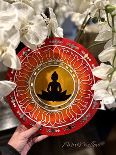 a person holding a red plate with a buddha image on it and white flowers in the background