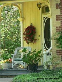 a yellow house with a chair and flowers on the front porch, surrounded by greenery