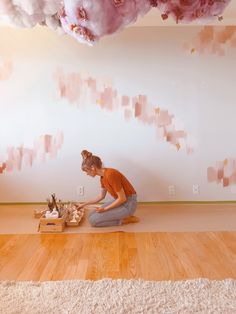 a woman sitting on the floor in front of a wall with pink clouds hanging from it