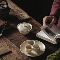 a person sitting at a table reading a book with cups and saucers on it