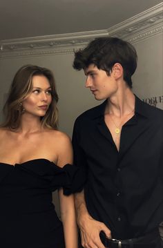 two people standing next to each other in front of a white wall and one person wearing a black dress