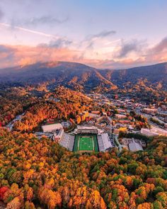 an aerial view of a football field surrounded by trees in the fall with mountains in the background