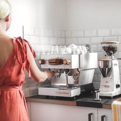 a woman in an orange dress is looking at the coffee machine on the counter with two espresso machines behind her