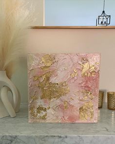 a pink and gold painting sitting on top of a marble counter next to vases