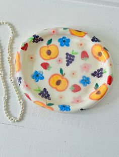 a white plate with fruit and flowers on it next to a beaded necklace hanging from the wall