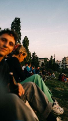 people are sitting on the grass and watching something in the distance with buildings in the background