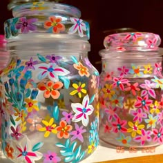 three jars with painted flowers on them are sitting on a counter top, one is clear and the other is bright