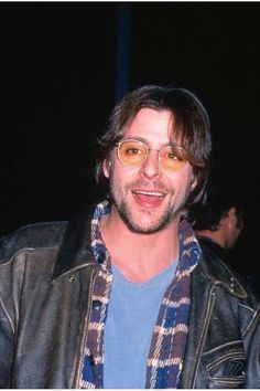 a man wearing glasses and a plaid scarf smiles at the camera while standing in front of a crowd